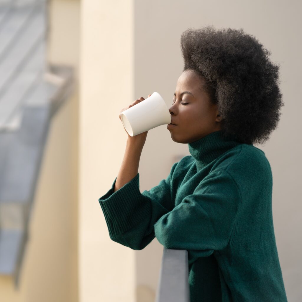 African American biracial woman enjoying a cup of tea or coffee, standing on balcony in the morning
